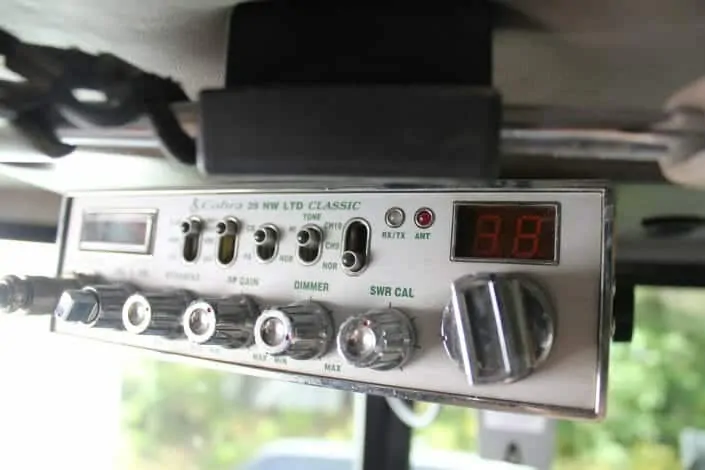A photo of an older CB radio. It is attached to the ceiling of a truck's cabin. It is a silver box with many dials, and a small red display. 