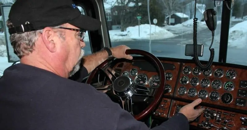 A photo of truck driver of a big rig, from behind the driver's shoulder. The truck's instrument panel and steering wheel can clearly be seen. 