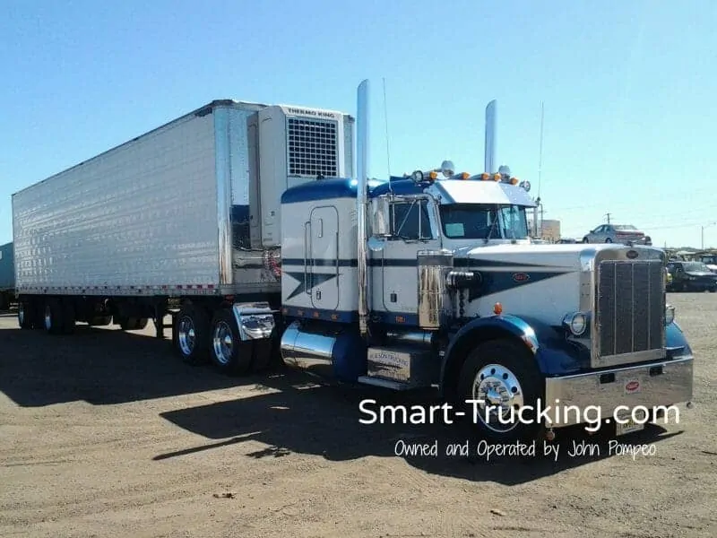 A photo of a 1987 long-nose Peterbilt 359-numbered truck. The tractor is colored white, with blue detailing. There's a large white trailer attached. 