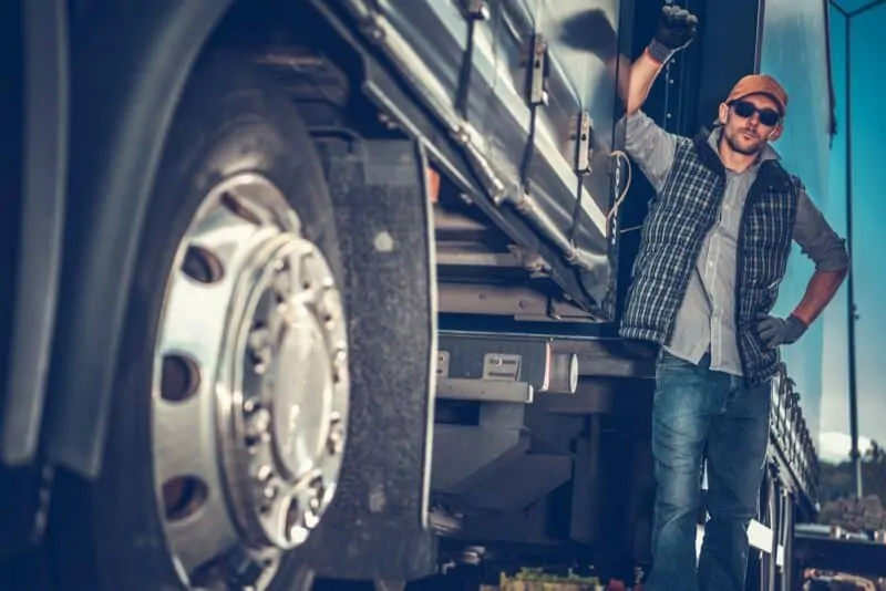 A photo of a truck driver leaning on big rig truck. The truck's wheel can be seen closely in the foreground. 